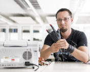 Inmitten der hellen, modernen Arbeitsräume der Hochschule Coburg sitzt eine Person mit Brille und Bart an einem Tisch in einem Labor und hält eine Handprothese in der Hand. Umgeben von elektronischen Geräten verkörpert sie Innovation und Fachwissen in dieser hochmodernen Umgebung.