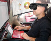Eine Person mit Virtual-Reality-Brille sitzt an einem Laptop mit roter Tastatur und ist in eine High-Tech-Multitasking-Arbeitsumgebung eingetaucht. Auf mehreren Computerbildschirmen werden Grafiken und Dokumente angezeigt, was den Innovationsgeist der Hochschule Coburg widerspiegelt.