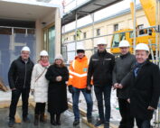Eine Gruppe von sieben Personen mit Bauhelmen und Winterkleidung steht auf einer Baustelle der Hochschule Coburg. Sie lächeln und scheinen für das Foto zu posieren, im Hintergrund sind Baumaschinen und Gerüste zu sehen.