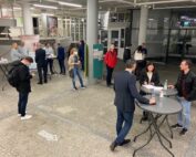 In einem modernen Innenraum der Hochschule Coburg haben sich Menschen versammelt. Sie sind leger gekleidet, stehen an hohen Tischen, manche unterhalten sich. Im Hintergrund ist ein Werbeplakat zu sehen. Der Boden ist grau und der Raum gut beleuchtet.