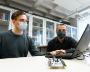Zwei Männer mit Masken arbeiten in einem Labor der Hochschule Coburg an einem Laptop. Sie konzentrieren sich konzentriert auf ein kleines elektronisches Gerät auf dem Tisch, umgeben von Regalen mit Ordnern und geschlossenen Schränken im Hintergrund.