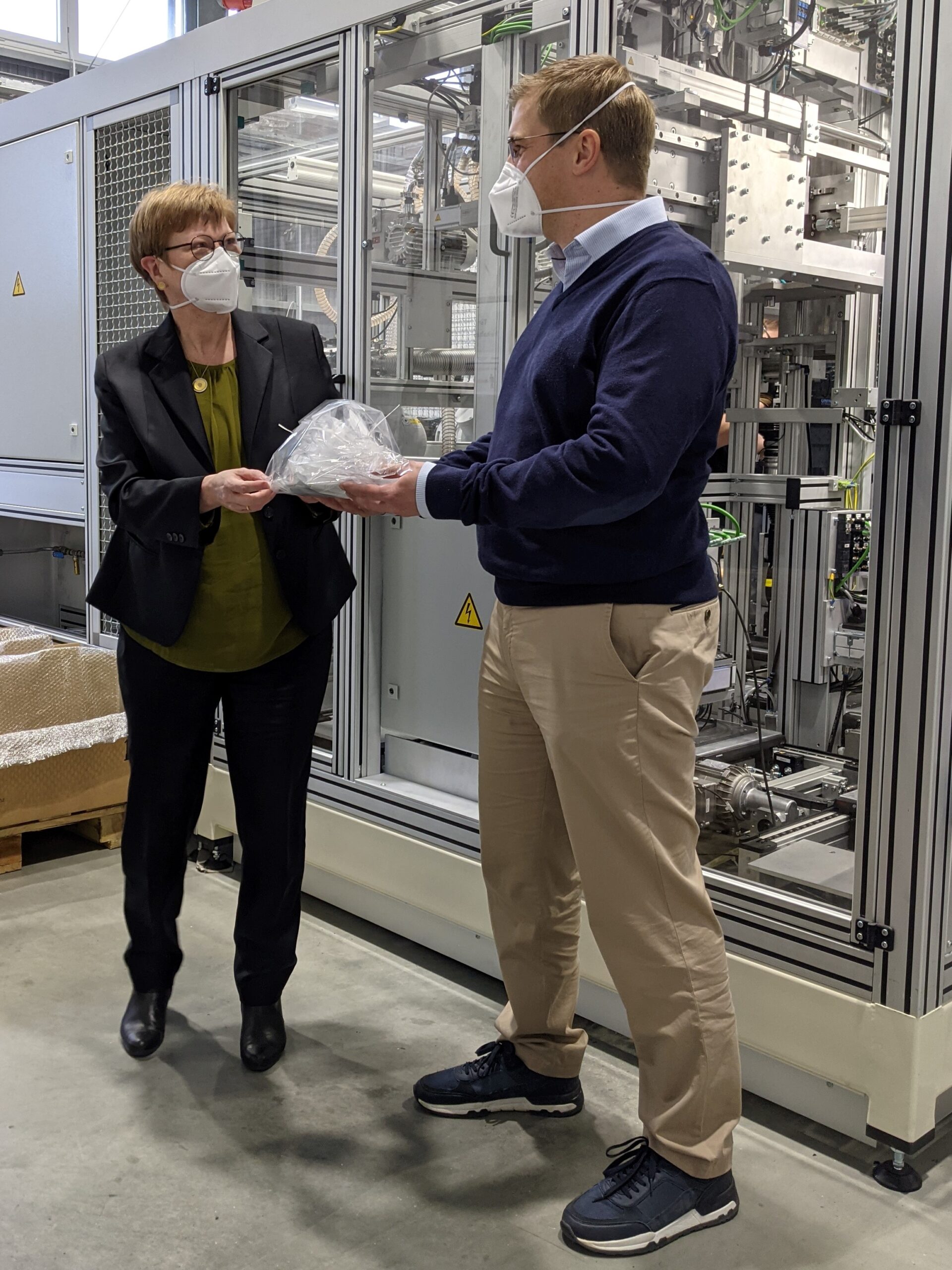 Zwei Menschen mit Masken stehen in einer Produktionsstätte der Hochschule Coburg. Einer reicht dem anderen einen Sack mit weißem Material, während sie vor Industriemaschinen stehen, an deren Seite sich Stapel mit Materialien befinden. Beide sind in legerer Business-Kleidung gekleidet und verkörpern eine Mischung aus Professionalität und Innovation.