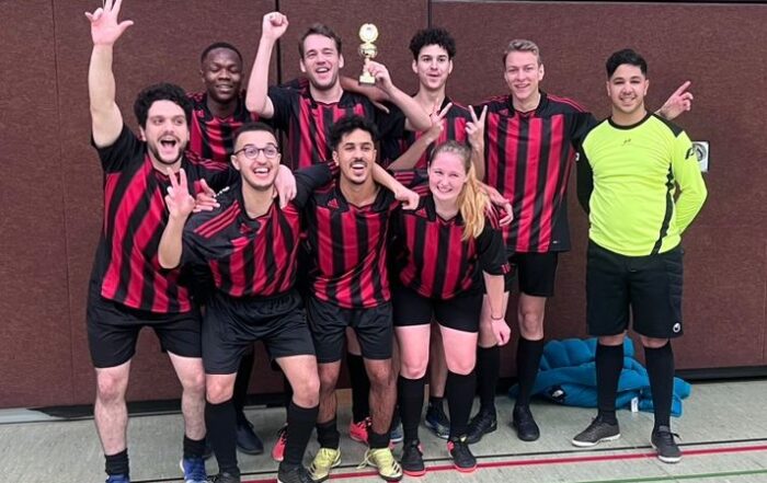Eine Mannschaft aus neun Fußballspielern der Hochschule Coburg in schwarz-rot gestreiften Trikots posiert gemeinsam im Innenbereich und hält einen kleinen Pokal in den Händen. Sie lächeln und machen feierliche Gesten.