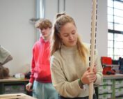 Eine junge Frau in einem beigen Pullover untersucht eine hohe Holzkonstruktion in der Werkstatt der Hochschule Coburg. Hinter ihr beobachtet ein junger Mann in einem roten Kapuzenpullover. Der Raum ist voller Werkzeuge und Materialien auf Regalen und Tischen, alles in natürliches Licht getaucht, das durch die Fenster fällt.