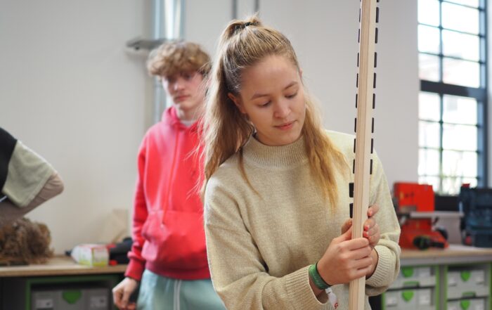 Eine junge Frau in einem beigen Pullover untersucht eine hohe Holzkonstruktion in der Werkstatt der Hochschule Coburg. Hinter ihr beobachtet ein junger Mann in einem roten Kapuzenpullover. Der Raum ist voller Werkzeuge und Materialien auf Regalen und Tischen, alles in natürliches Licht getaucht, das durch die Fenster fällt.