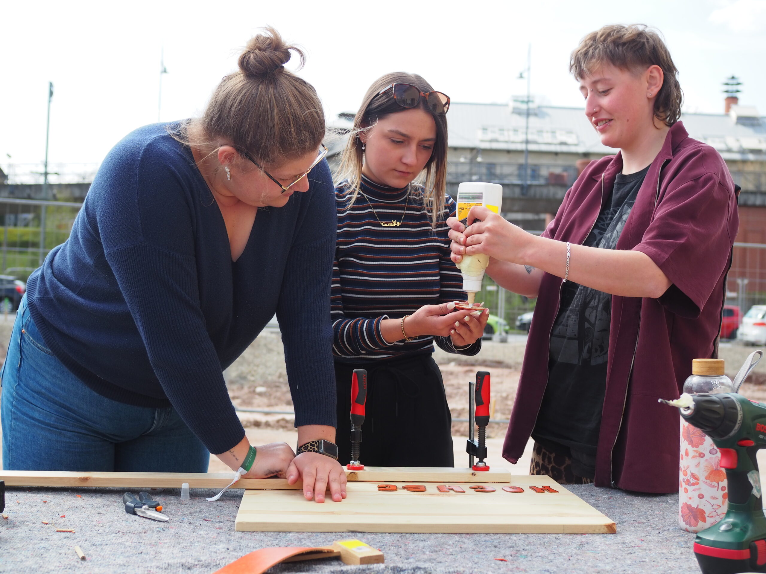 Drei Personen arbeiten gemeinsam an einem Holzbearbeitungsprojekt im Freien an der Hochschule Coburg. Einer hält eine Flasche Klebstoff, während die anderen assistieren. Der Tisch ist mit Werkzeugen und Holzstücken bedeckt. Sie wirken konzentriert und arbeiten unter einem bewölkten Himmel zusammen.