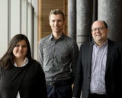 Drei Personen stehen in einem Innenraum an großen Fenstern der Hochschule Coburg. Eine Frau links trägt einen schwarzen Pullover, ein Mann in der Mitte ein gemustertes Hemd und ein Mann rechts einen Anzug. Sie alle lächeln in die Kamera.