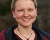 Eine Person mit kurzen Haaren lächelt herzlich, trägt ein kastanienbraunes Hemd und eine dunkle Halskette unter einer offenen Jacke und verkörpert den jugendlichen Geist der Hochschule Coburg. Die verschwommene Naturkulisse im Hintergrund verleiht der Szene eine heitere Note.