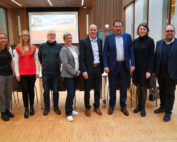 Eine Gruppe von acht Personen steht auf einem Holzboden in einem modernen Raum der Hochschule Coburg. Sie sind in legerer Business-Kleidung gekleidet, im Hintergrund ist ein Präsentationsbildschirm zu sehen.