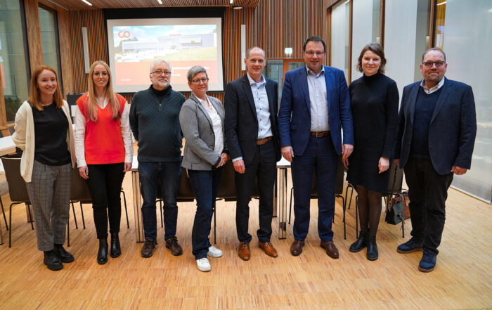 Eine Gruppe von acht Personen steht auf einem Holzboden in einem modernen Raum der Hochschule Coburg. Sie sind in legerer Business-Kleidung gekleidet, im Hintergrund ist ein Präsentationsbildschirm zu sehen.