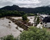 Ein über die Ufer getretener Fluss mit Geröll überflutet die Szene und überschwemmt Gebäude in einem kleinen Dorf in der Nähe der Hochschule Coburg. Saftig grüne Hügel bilden den Hintergrund, während der bewölkte Himmel auf heftige Regenfälle der letzten Zeit hindeutet.