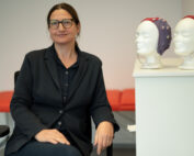 Eine Person im schwarzen Anzug sitzt in einer büroähnlichen Umgebung an der Hochschule Coburg. In der Nähe stehen zwei mit bunten EEG-Kappen geschmückte Puppenköpfe. Im Hintergrund sorgt eine rote Bank für einen Farbtupfer in der Szene.