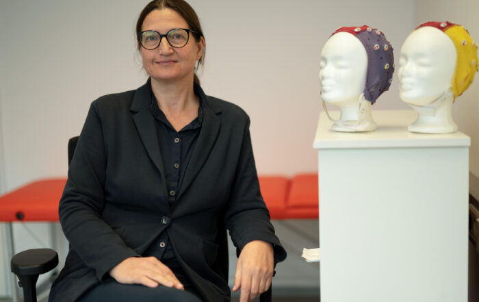 Eine Person im schwarzen Anzug sitzt in einer büroähnlichen Umgebung an der Hochschule Coburg. In der Nähe stehen zwei mit bunten EEG-Kappen geschmückte Puppenköpfe. Im Hintergrund sorgt eine rote Bank für einen Farbtupfer in der Szene.