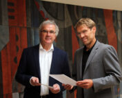 Zwei Männer in Anzügen stehen in einem Gebäude der Hochschule Coburg, halten Dokumente in den Händen und lächeln. Sie stehen vor einem farbenfrohen abstrakten Wandgemälde mit geometrischen Mustern.