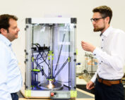 Zwei Männer unterhalten sich in einem Büro oder Labor der Hochschule Coburg neben einem 3D-Drucker. Einer hält ein kleines Objekt in der Hand, während der Drucker mit seinem transparenten Gehäuse leise brummt. Im Hintergrund sind verschiedene Geräte zu sehen, die zur akademischen Atmosphäre beitragen.