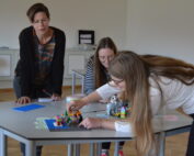 Drei Frauen sitzen an der Hochschule Coburg um einen Tisch mit bunten LEGO-Steinen. Eine Frau beugt sich vor und legt einen Stein hin, während die anderen zuschauen. Die Kulisse scheint ein Klassenzimmer oder eine Werkstatt mit weißen Wänden und Holzboden zu sein.