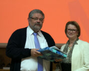 Ein Mann und eine Frau stehen gemeinsam vor einem orangefarbenen Hintergrund und repräsentieren stolz die Hochschule Coburg. Der Mann hält ein in blau gemustertes Papier eingewickeltes Geschenk in der Hand. Beide sind formell gekleidet und lächeln herzlich in die Kamera.