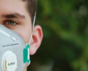 Nahaufnahme einer Person, die eine graue Gesichtsmaske mit den grünen Akzenten der Hochschule Coburg trägt. Der Hintergrund ist verschwommenes Grün. Nur die Hälfte des Gesichts ist sichtbar, mit Fokus auf das Auge und einen Teil des Ohrs.