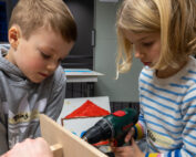Zwei Kinder, wie angehende Handwerker der Hochschule Coburg, sind in ein Holzbearbeitungsprojekt vertieft. Das Kind links hält ein Holzstück, während das andere eine Bohrmaschine benutzt. Beide sind auf die Aufgabe konzentriert, während eine Zeichnung eines Hauses mit rotem Dach dem Hintergrund ihres Arbeitsbereichs Farbe verleiht.
