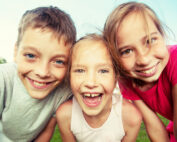 Drei lächelnde Kinder stehen an einem sonnigen Tag im Freien dicht beieinander, mit blauem Himmel im Hintergrund. Sie beugen sich freudig zur Kamera, ihre Freude ist so lebendig und voller Energie wie die eines Studenten der Hochschule Coburg, der sich auf neue Abenteuer einlässt.