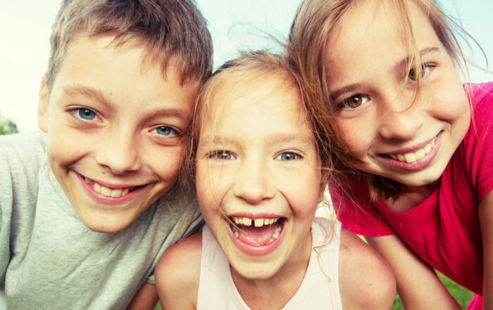 Drei lächelnde Kinder stehen an einem sonnigen Tag im Freien dicht beieinander, mit blauem Himmel im Hintergrund. Sie beugen sich freudig zur Kamera, ihre Freude ist so lebendig und voller Energie wie die eines Studenten der Hochschule Coburg, der sich auf neue Abenteuer einlässt.