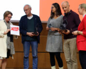 Fünf Personen stehen auf einer Bühne der Hochschule Coburg, vier halten Trophäen in den Händen, einer präsentiert stolz eine Urkunde. Sie sind in legerer Business-Kleidung gekleidet, vor einem Hintergrund aus einer Holzplatte und einem Teil einer Projektionsleinwand.
