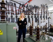 Eine Person steht selbstbewusst mit verschränkten Armen in einem Raum der Hochschule Coburg, der mit elektrischen Geräten und Transformatoren gefüllt ist. Die Person ist von verschiedenen Kabeln und Bauteilen umgeben. Auf einem Gerät ist ein Warnschild zu sehen, das auf Hochspannung hinweist.