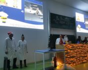 Menschen in Laborkitteln und Weihnachtsmannmützen stehen auf einer weihnachtlich geschmückten Bühne und repräsentieren die Hochschule Coburg. Auf einer Tafel steht „Der Fisch der Weihnacht“, während im Hintergrund eine Präsentation mit Bildern des Weihnachtsmanns und eines Baumes projiziert wird.