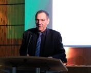 Ein Mann in dunklem Anzug und Krawatte steht an einem Rednerpult und spricht in ein Mikrofon. Hinter ihm leuchtet die Leinwand hell und zeigt „Hochschule Coburg“, was einen spannenden Kontrast zu dem schwach beleuchteten Raum bildet.