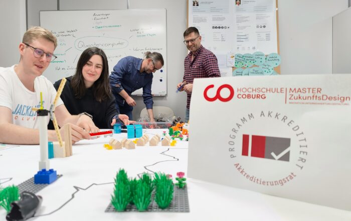 Eine Gruppe von vier Personen sitzt um einen mit LEGO-Steinen und Papieren beladenen Tisch und arbeitet intensiv zusammen. Das Bild wird dominiert von einem prominent platzierten Poster des Masterstudiengangs ZukunftsDesign der Hochschule Coburg, das durch seine auffällige Präsenz die Aufmerksamkeit auf sich zieht.