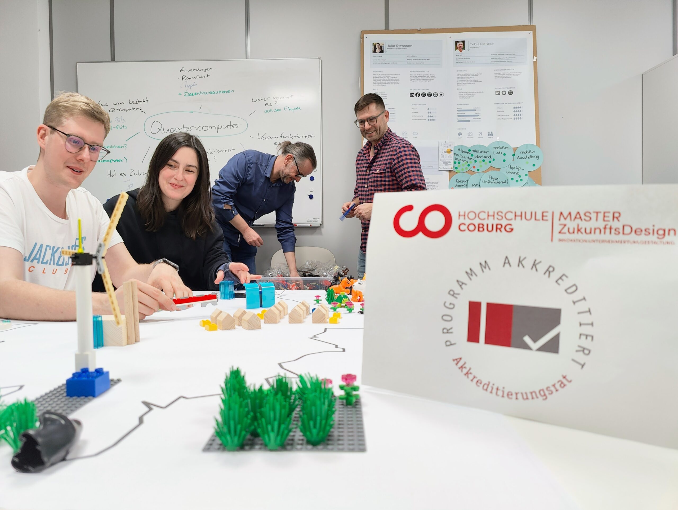 Eine Gruppe von vier Personen sitzt um einen mit LEGO-Steinen und Papieren beladenen Tisch und arbeitet intensiv zusammen. Das Bild wird dominiert von einem prominent platzierten Poster des Masterstudiengangs ZukunftsDesign der Hochschule Coburg, das durch seine auffällige Präsenz die Aufmerksamkeit auf sich zieht.