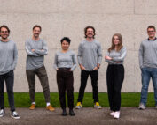 Sechs Menschen in Freizeitkleidung stehen vor einer schlichten Betonwand an der Hochschule Coburg, blicken in die Kamera und lächeln.