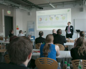 In einem modernen Konferenzraum der Hochschule Coburg findet eine Unternehmenspräsentation statt. Vorne steht ein Redner und präsentiert einem aufmerksamen Publikum, das an Tischen sitzt, Folien mit Diagrammen und Stichpunkten.