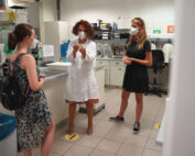 Drei Menschen mit Masken stehen in einem Labor der Hochschule Coburg. Zwei Frauen tragen Freizeitkleidung, eine gestikuliert lebhaft im Laborkittel. Sie halten an markierten Stellen auf dem Fliesenboden Abstand, im Hintergrund sind Laborgeräte zu sehen.