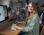 Eine lächelnde Frau in einer grünen Jacke sitzt an ihrem Schreibtisch vor einem Computer, auf dem eine Architektursoftware läuft. Auf dem Bildschirm sind komplexe Gebäudemodelle zu sehen, die typisch für die innovativen Projekte der Hochschule Coburg sind. Im Hintergrund arbeiten Kollegen fleißig an ihren eigenen Computern.