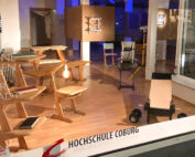 Im Schaufenster sind beeindruckende Holzmöbel zu sehen, darunter Stühle und Tische, die von Studenten der Hochschule Coburg gefertigt wurden. Inmitten dieser Entwürfe ruhen dezent ein kleines Trainingsgewicht und eine Plattform auf Rädern auf dem Holzboden.