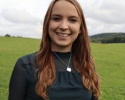 Eine Person mit langen braunen Haaren und einer Halskette steht lächelnd auf einer grünen Wiese unter dem bewölkten Himmel in der Nähe der Hochschule Coburg.