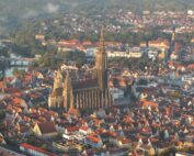 Eine Luftaufnahme einer Stadt zeigt eine markante gotische Kathedrale mit einem hohen Turm in der Mitte. Die Umgebung, in deren Nähe sich die Hochschule Coburg befindet, zeichnet sich durch zahlreiche Gebäude mit roten Dächern, Straßen und Grünflächen aus, alles vor der Kulisse eines gewundenen Flusses.