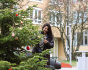 Vor der Hochschule Coburg schmückt eine Frau einen Weihnachtsbaum mit Ornamenten. Ihr Lächeln strahlt unter ihrer kuscheligen Winterjacke. Der Baum glänzt mit roten und goldenen Dekorationen vor einer Kulisse aus blattlosen Bäumen und den Fenstern des Gebäudes.