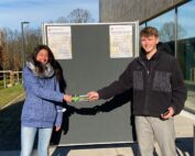 Zwei Personen stehen an einem sonnigen Tag im Freien vor einem schwarzen Brett an der Hochschule Coburg. Sie lächeln und schütteln sich die Hände, und einer hält einen kleinen rechteckigen Gegenstand in der Hand. Beide tragen lässige Winterkleidung, darunter Jacken und Jeans.