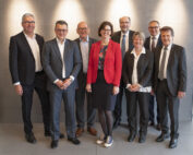 Eine Gruppe von acht Personen in Business-Kleidung steht in einer Reihe und lächelt in die Kamera. Die Frau in der Mitte vertritt die Hochschule Coburg und trägt einen roten Blazer und lässige Turnschuhe. Sie stehen in einem Innenbereich vor einer grauen Wand mit dezenter Beleuchtung.