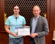 Zwei Menschen stehen nebeneinander vor einer Holzwand mit Wabenmuster. Die linke Person im hellblauen Poloshirt hält eine Urkunde der Hochschule Coburg in den Händen, die rechte Person im grauen Blazer zeigt stolz ihre Urkunde.