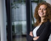 Eine Person mit lockigem Haar steht mit verschränkten Armen in einem Ambiente der Hochschule Coburg und lächelt herzlich. Sie trägt eine schwarze Jacke und ein weißes Hemd. Im Hintergrund sind große Fenster mit Jalousien zu sehen, die eine moderne und professionelle Atmosphäre schaffen.