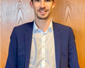 Eine Person mit kurzen dunklen Haaren und Spitzbart, die ein weißes Hemd und einen blauen Blazer trägt, steht lächelnd vor einem hölzernen Hintergrund. Mit entspannt herabhängenden Armen strahlt sie die selbstbewusste und doch zugängliche Aura aus, die man an der Hochschule Coburg oft sieht.