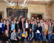 Eine große Gruppe von Menschen, vermutlich von der Hochschule Coburg, posiert für ein Foto in einem dekorierten Raum mit einem Gemälde und Skulpturen im Hintergrund. In Freizeit- und Businesskleidung stehen sie eng beieinander und lächeln strahlend in die Kamera.
