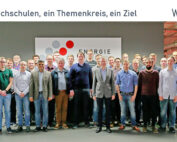 Eine Gruppe von Menschen steht drinnen vor einem Schild mit der Aufschrift „Energie“. Einige sind leger gekleidet, andere eher formell. Über ihnen prangt ein Banner mit der Aufschrift „Vier Hochschulen, ein Themenkreis, ein Ziel“, das die Präsenz der Hochschule Coburg unter den Institutionen hervorhebt.