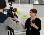 Eine Frau mit Brille steht vor einem Whiteboard an der Hochschule Coburg, spricht angeregt und gestikuliert mit ihren Händen. Währenddessen hält ein Mann mit einer Beaniemütze den Moment auf Video fest. Die akademische Atmosphäre ist lebendig und konzentriert.