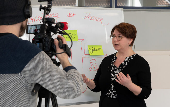Eine Frau mit Brille steht vor einem Whiteboard an der Hochschule Coburg, spricht angeregt und gestikuliert mit ihren Händen. Währenddessen hält ein Mann mit einer Beaniemütze den Moment auf Video fest. Die akademische Atmosphäre ist lebendig und konzentriert.
