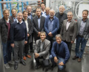 Eine Gruppe von vierzehn Männern in legerer und halbformeller Kleidung steht und kniet in einem Raum der Hochschule Coburg, im Hintergrund sind Industrieanlagen und Metallgeländer zu sehen. Sie scheinen für ein Gruppenfoto zu posieren.