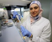 Eine Wissenschaftlerin der Hochschule Coburg, die Hijab, einen weißen Laborkittel und blaue Handschuhe trägt, benutzt im Labor eine Pipette. Im Hintergrund hängen Regale mit Geräten und Laborkitteln, während sie in die Kamera lächelt.